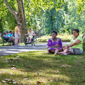 Two women sitting on the grass, smiling and talking, with a person in a wheelchair on a path in the background.