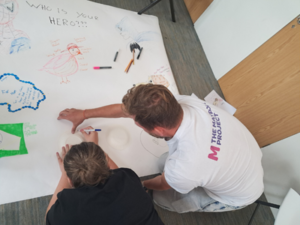 Overhead view of two people sitting on the floor collaborating on a large white poster. One person traces a paper plate while the other person from the Maypole Project assists. The poster is titled “WHO IS YOUR HERO???” and features hand-drawn illustrations, including a bird with descriptive labels and superhero figures.